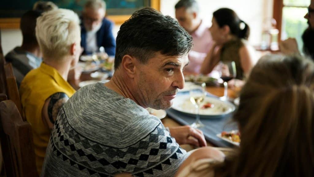 A serious man in a patterned sweater is seated at a table surrounded by other people.