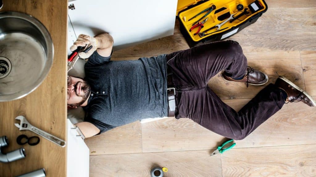 A man is lying on his back on a wooden floor, working under a kitchen sink with tools nearby.