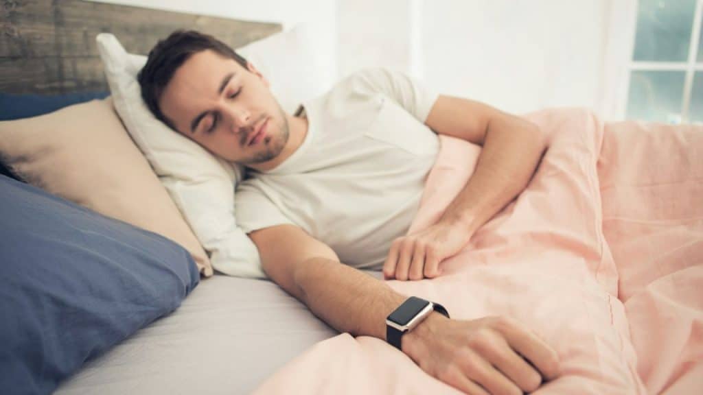 A man wearing a fitness tracker is sleeping in a bed under a light pink blanket.