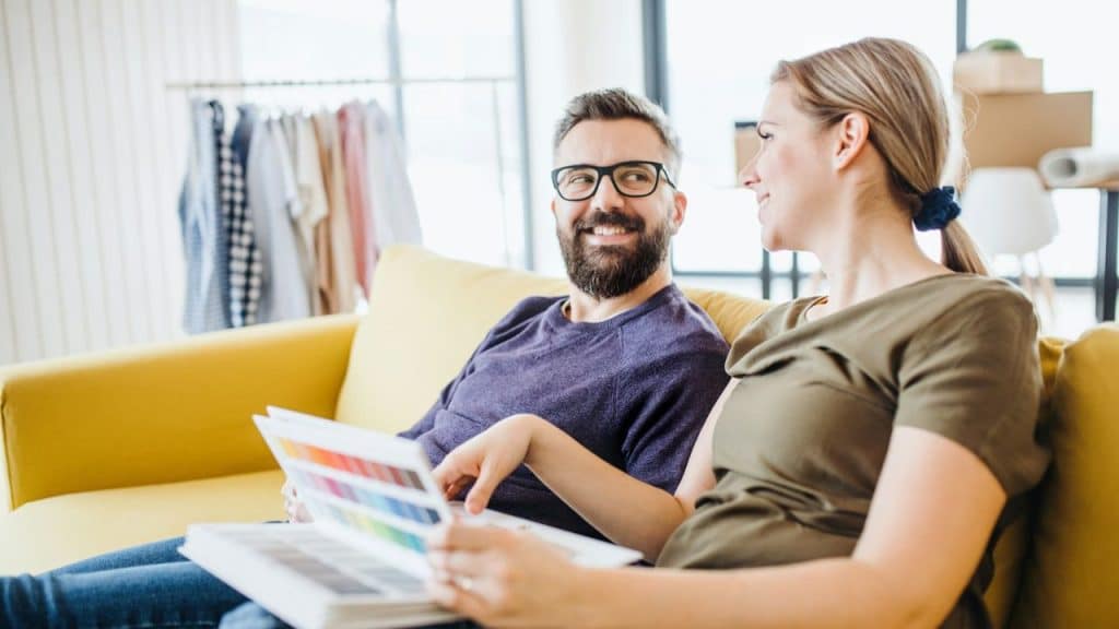 A happy couple sits on a yellow couch looking at paint color swatches.