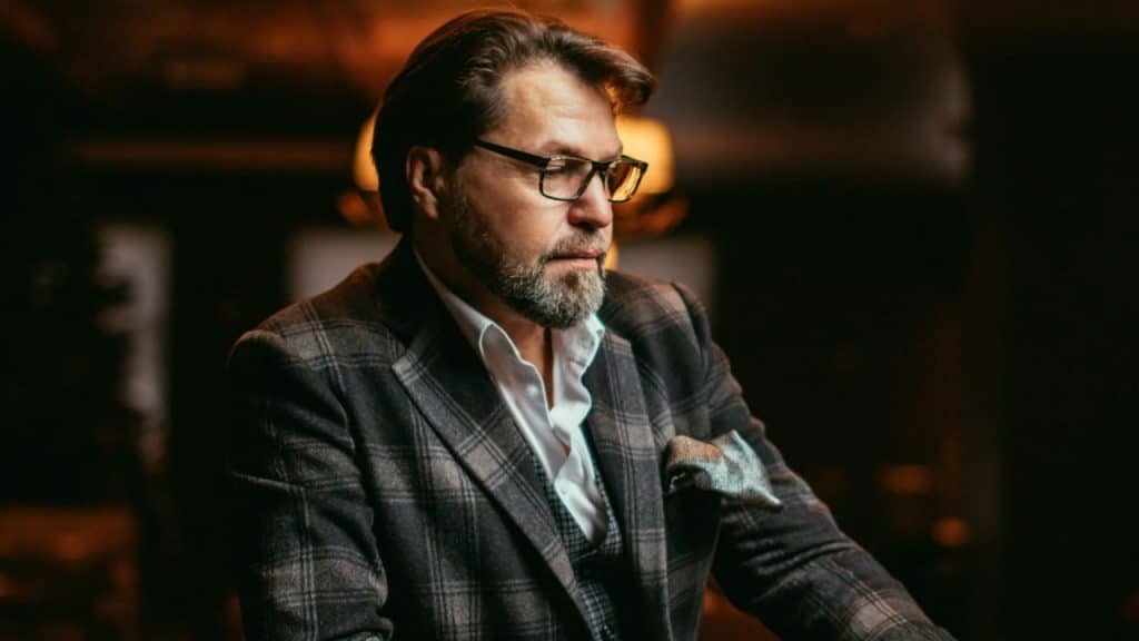 A bearded man in a plaid sport coat and glasses looks down in a dimly lit room.
