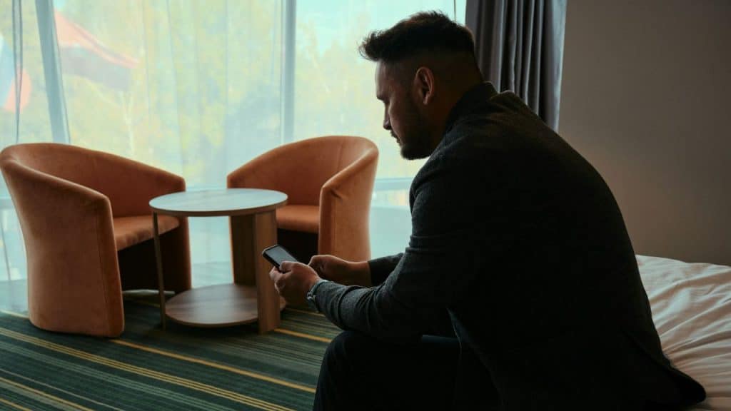 A man in a dark suit sits on a bed, looking at his phone, with two orange chairs and a table in the background.