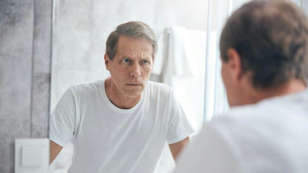 A serious-looking man in a white t-shirt looks intently at his reflection in a bathroom mirror.