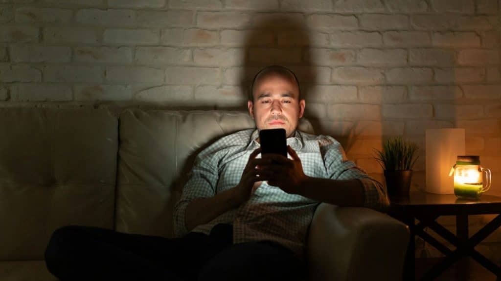 A bald man sits on a sofa in a dimly lit room, looking at his glowing cell phone.