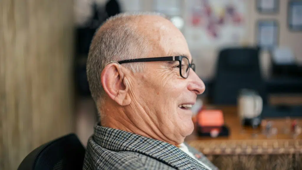 A close-up side profile of an older man with glasses smiling in an office setting.