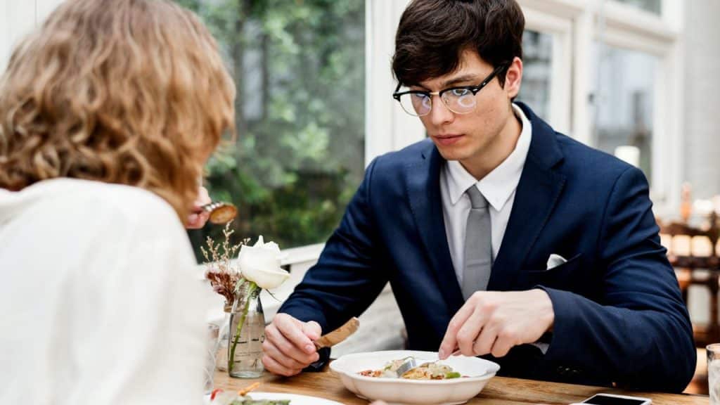 A man in a navy suit and glasses cuts food on a plate while having a meal with a woman.