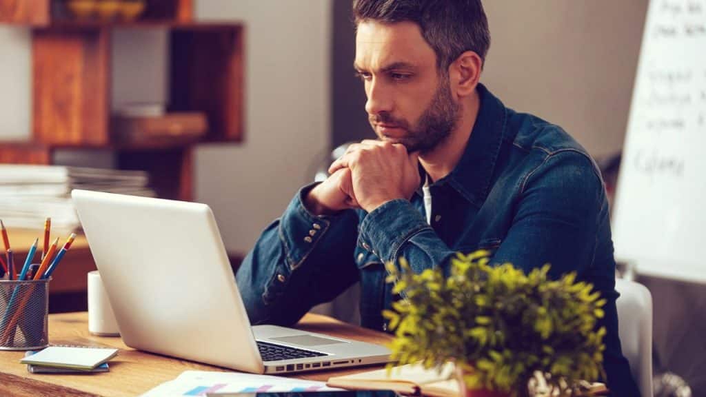 A focused man with a beard and denim shirt stares intently at a laptop screen in an office.