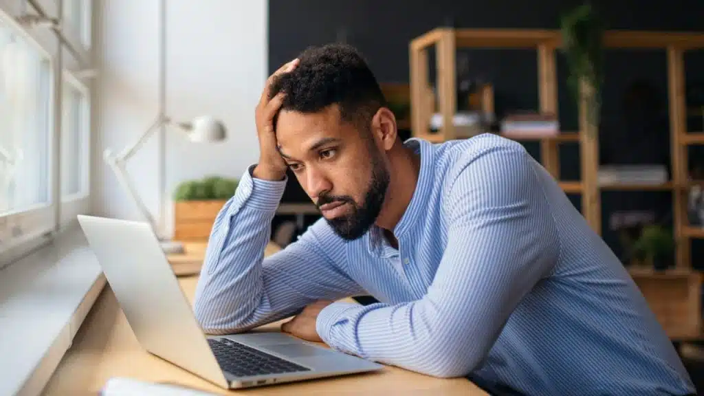 A stressed-looking man with a beard sits at a desk, hand on his head, staring at a laptop.