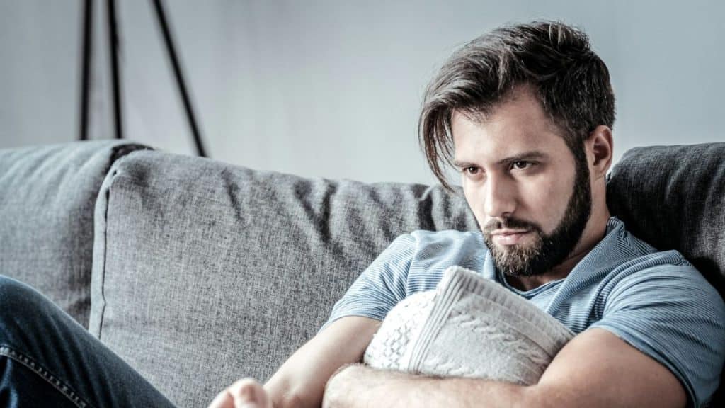 A bearded man sits on a gray sofa, holding a textured pillow and looking pensive.