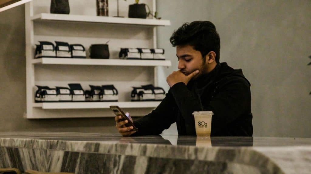 A young man with his hand on his chin looks at his phone while sitting at a marble counter with an iced coffee.