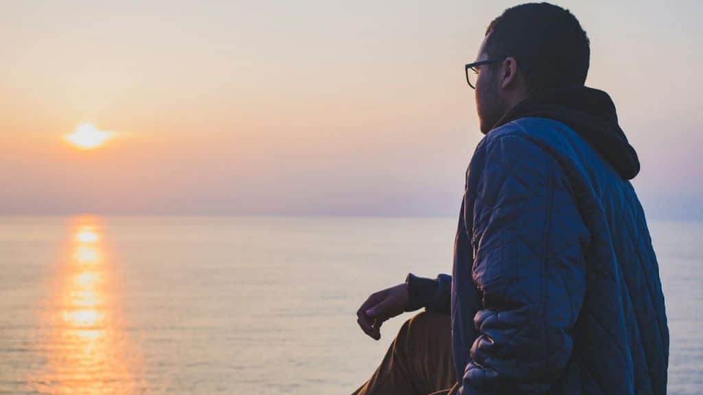 A man in a blue quilted jacket and glasses sits looking at a sunset over the sea.
