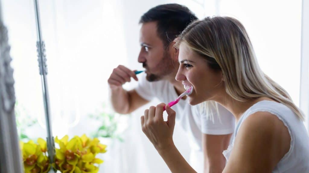 A woman and a man stand side-by-side, brushing their teeth in front of a mirror.