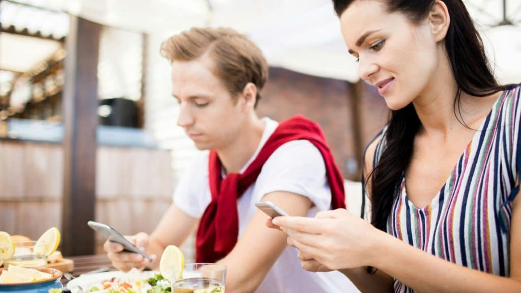 A woman and a man sit at a table with food, both looking at their mobile phones.
