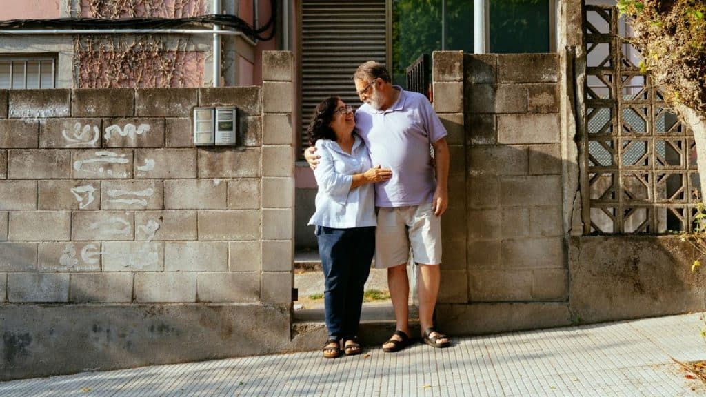 A middle-aged man and woman stand close, embracing, next to a concrete block wall.