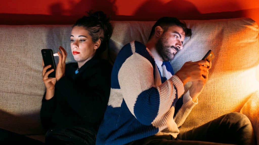 A woman and a man sit back-to-back on a couch, both looking at their mobile phones in a dimly lit room.