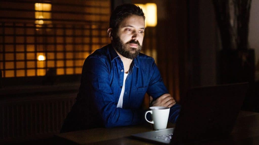 A bearded man in a denim shirt works on a laptop at night with a mug in front of him.