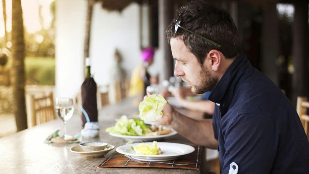 A man in a dark shirt sits at an outdoor table, looking at a piece of lettuce over a plate.