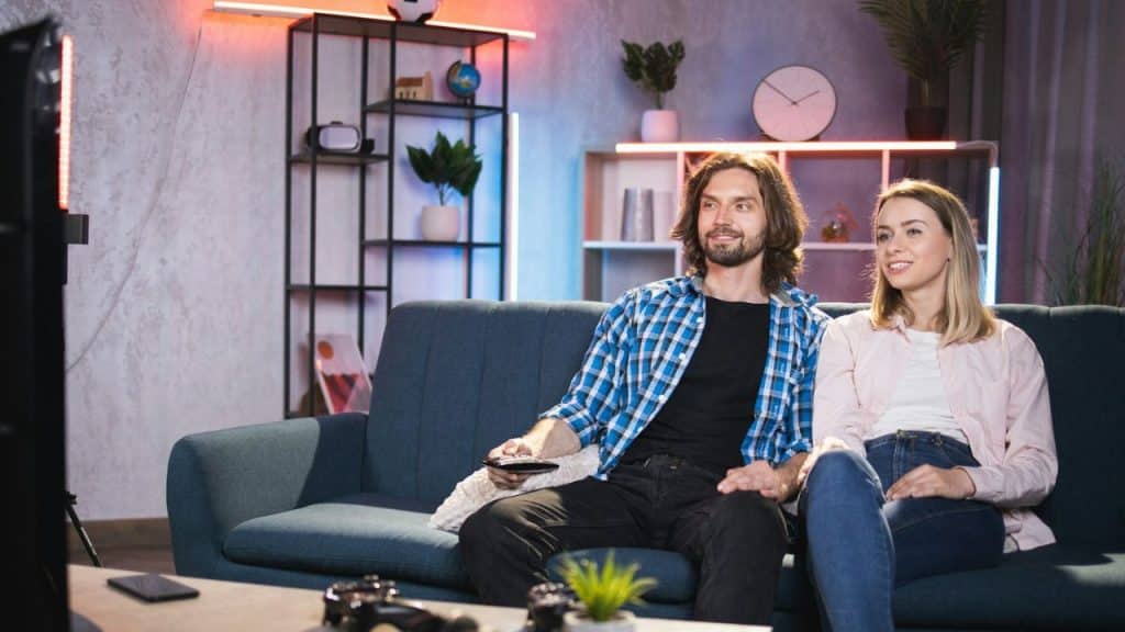 A man and woman sit together on a blue sofa watching a TV in a living room with LED lighting.