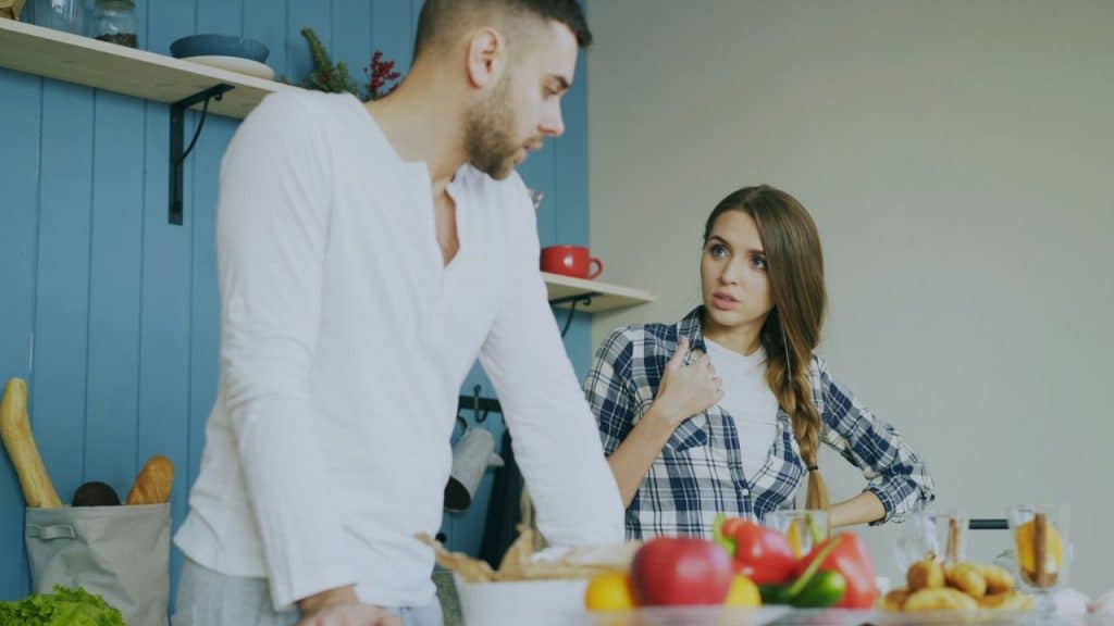 A man in a white shirt stands over a table, facing a woman in a plaid shirt looking concerned.