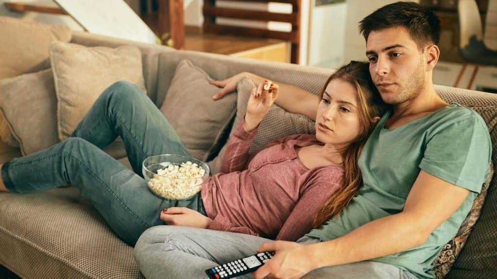A serious-looking couple watches TV on a couch, holding a bowl of popcorn and a remote control.