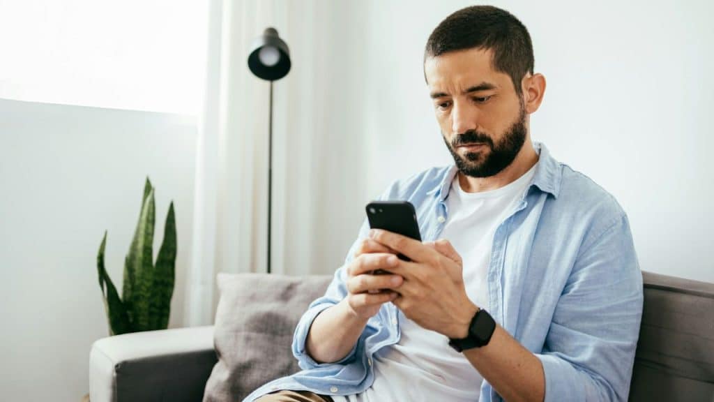 A bearded man in a light blue shirt sits on a sofa, intently looking at his mobile phone.