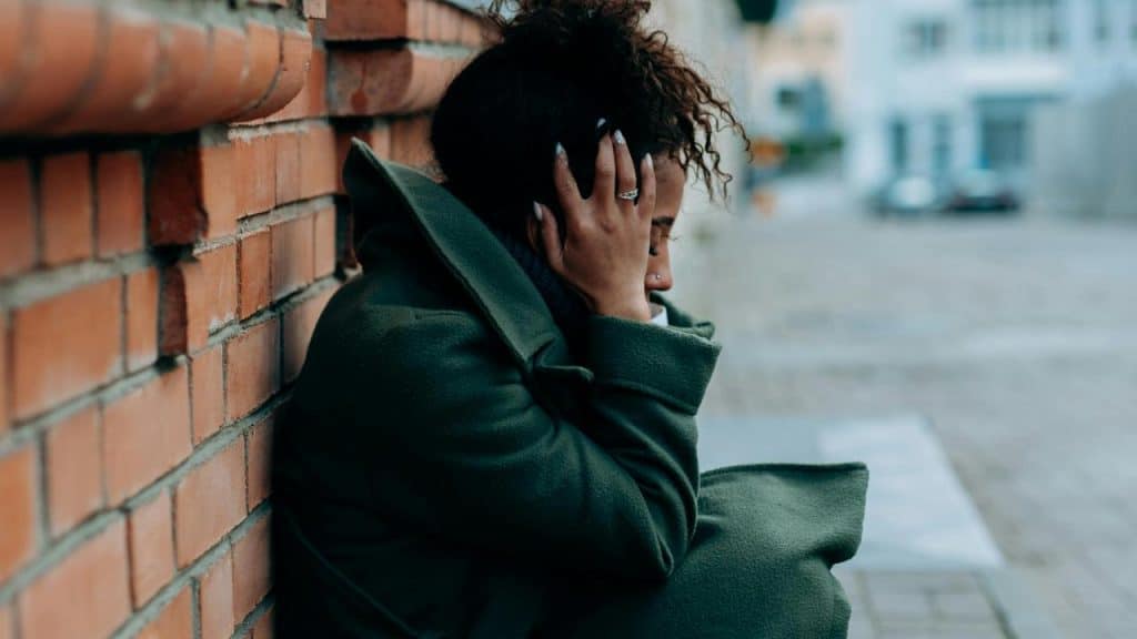 A person sitting by a brick wall with their hand on their head, appearing distressed.
