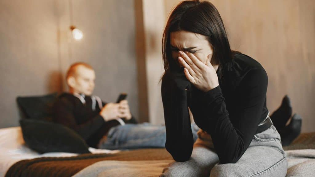 A person sitting on a bed and crying while another person sits in the background.