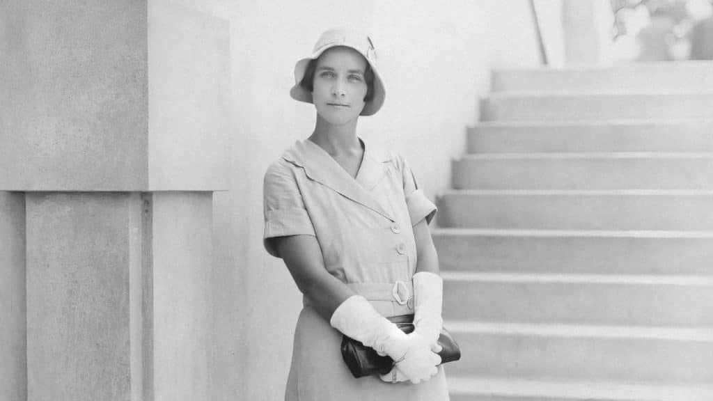 A black and white photo of a woman in a short-sleeved dress, cloche hat, and white gloves, standing by a stone staircase.