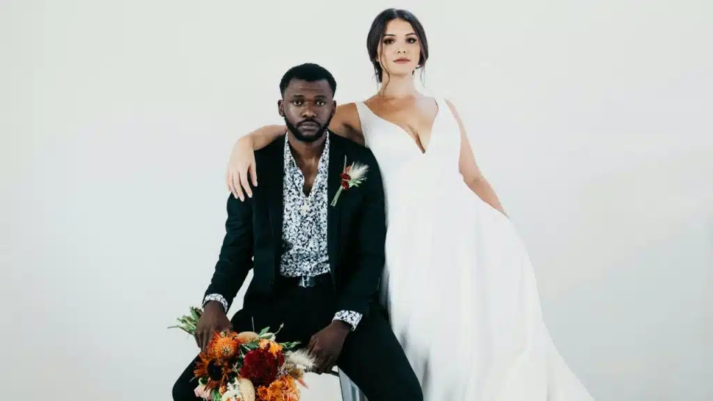 A wedding portrait of a Black man and a white woman, with the woman in a white dress standing and the man in a black suit seated holding a bouquet, against a white background.