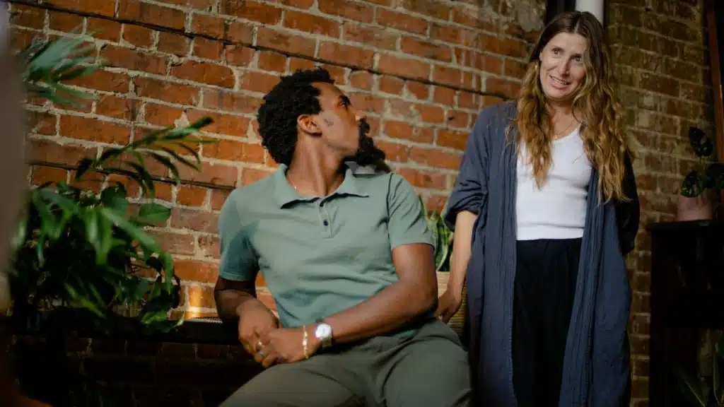 A Black man in a green polo shirt sits and looks toward a white woman with long wavy hair who is smiling at him, with a brick wall and plants in the background.