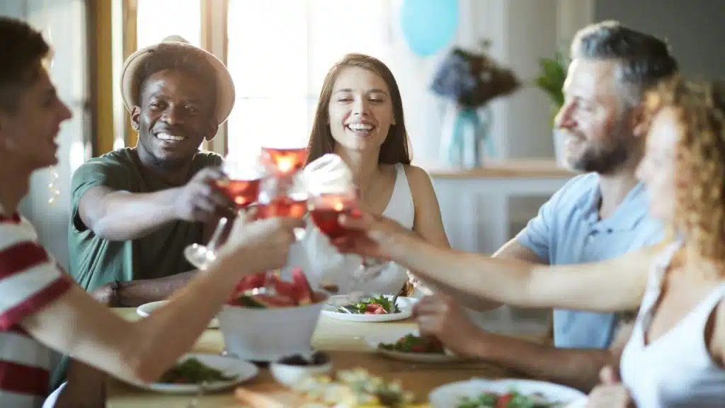 A diverse group of five people smiling and toasting with glasses of pink liquid around a dinner table with plates of salad.