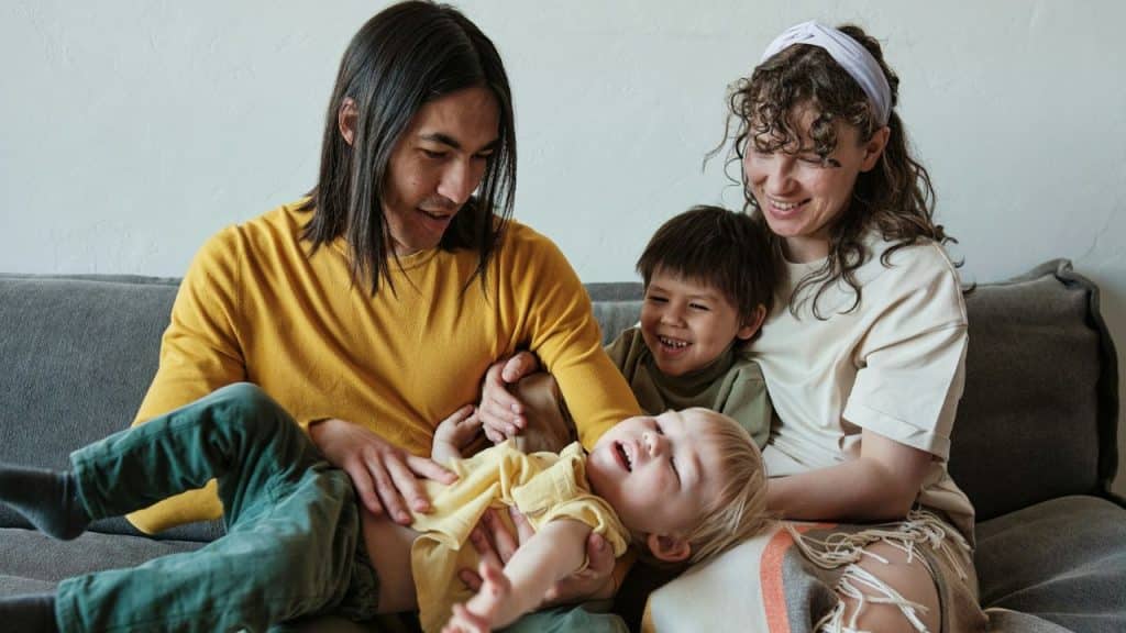 A diverse family of four is sitting on a couch, with a man in a yellow shirt holding a young child lying on his lap and a smiling woman with curly hair sitting next to an older boy.