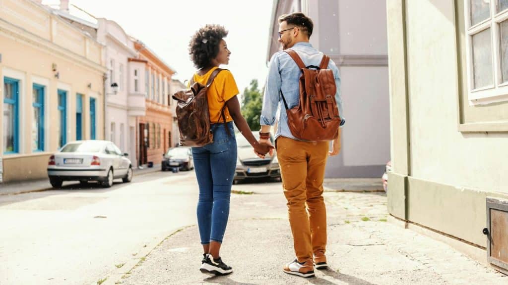An interracial couple is walking down a city street with brown leather backpacks, holding hands and looking at each other.