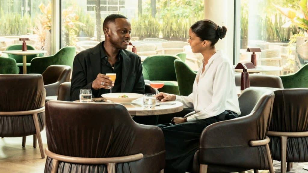 A Black couple is seated at a small table in a restaurant with large windows, eating and talking, with green velvet and brown leather chairs surrounding them.