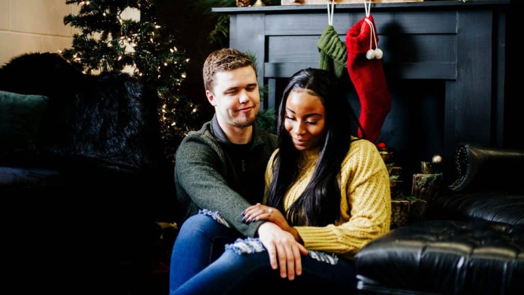 An interracial couple is seated on the floor in front of a fireplace with Christmas stockings and a decorated Christmas tree in the background.