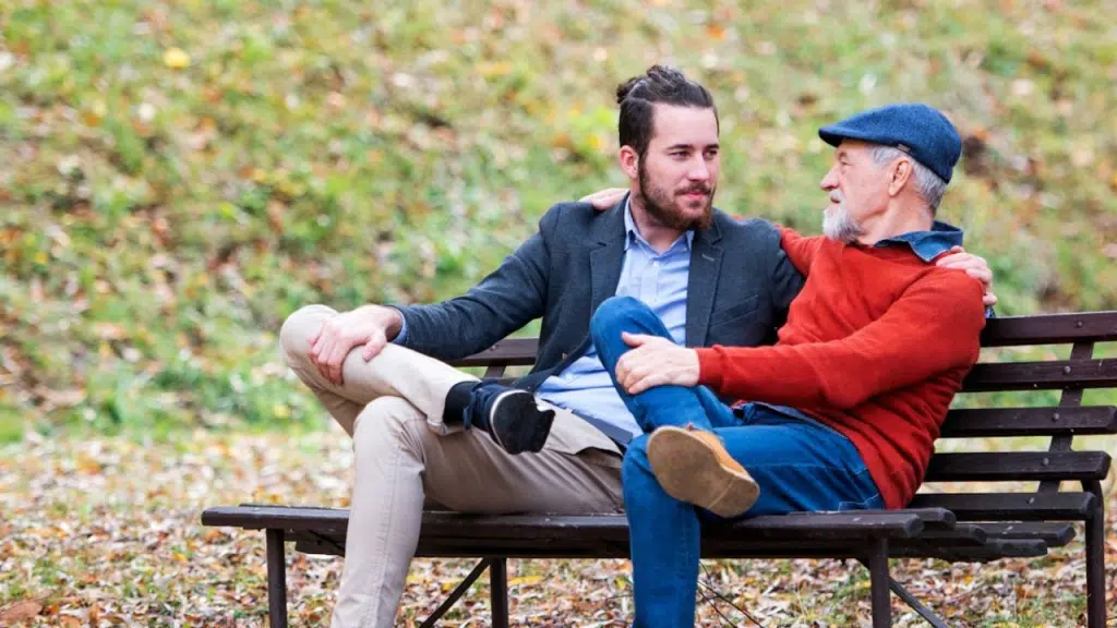 Two men of different ages sit on a park bench, talking and sharing an embrace.