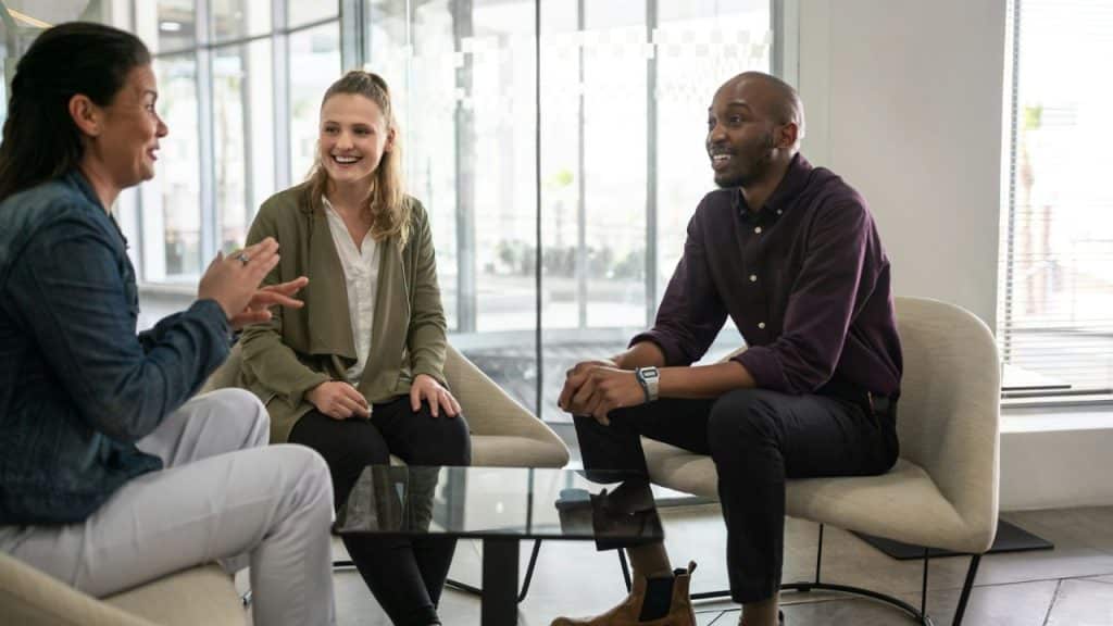 Three people are seated in modern chairs around a coffee table in a bright, glass-walled office space, talking and smiling.