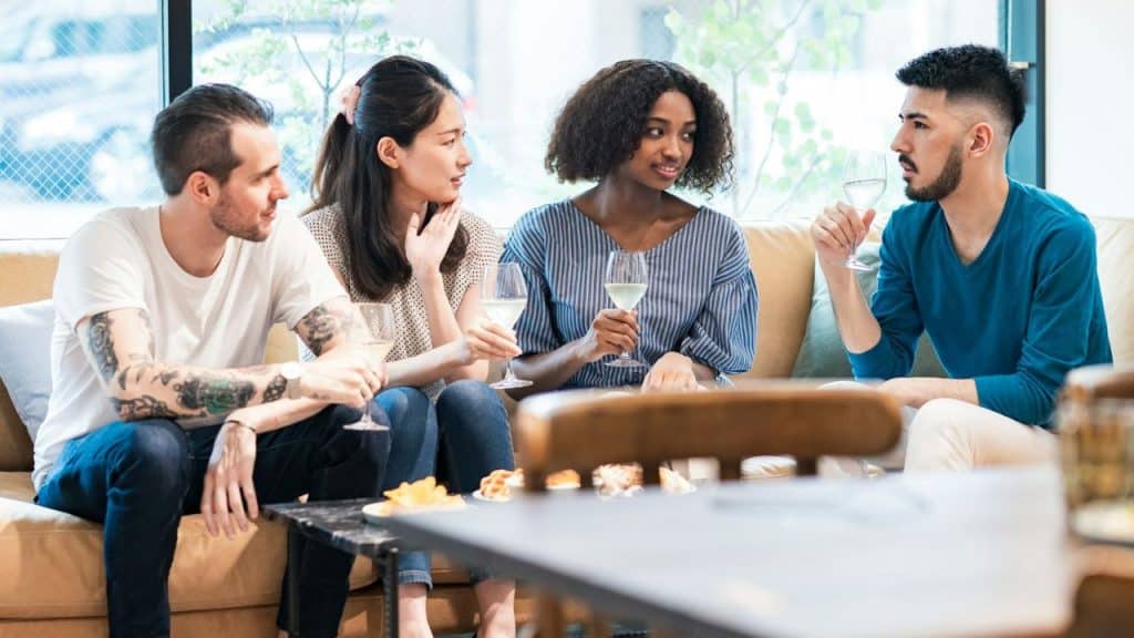 A diverse group of four friends is seated on a sofa, holding wine glasses and talking in a brightly lit room.