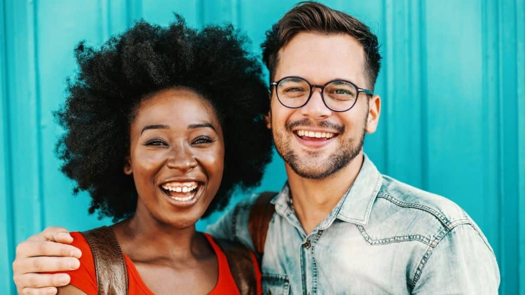 A smiling interracial couple is standing together in front of a turquoise wall, with the man's arm around the woman's shoulder.