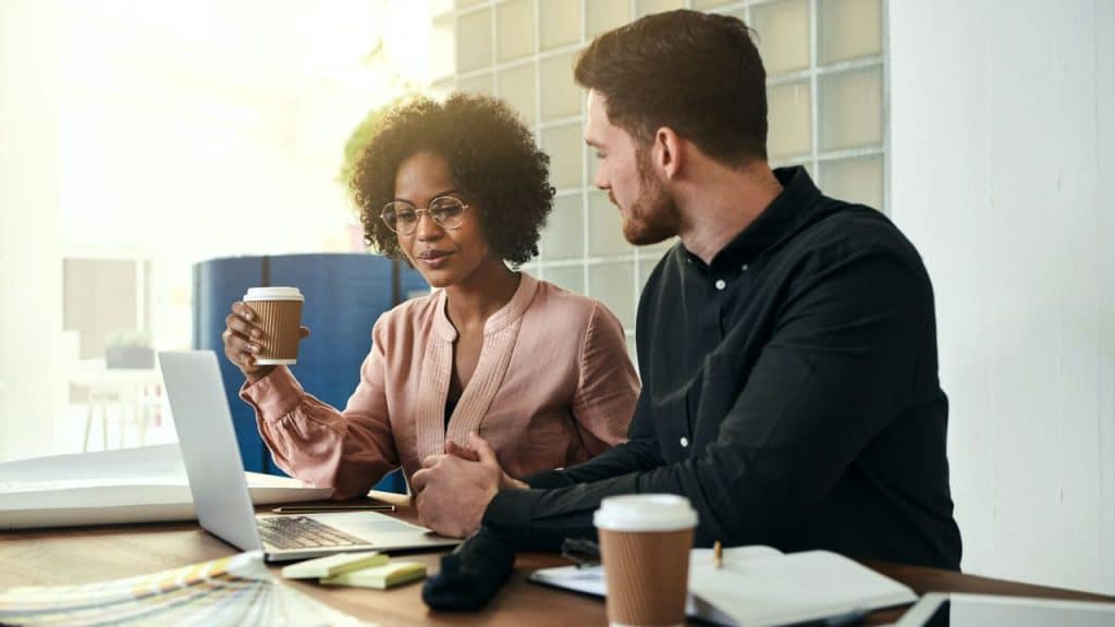 An interracial couple is seated at a table with a laptop and notebooks, with the Black woman holding a coffee cup and looking at the screen with a white man in a black shirt beside her.