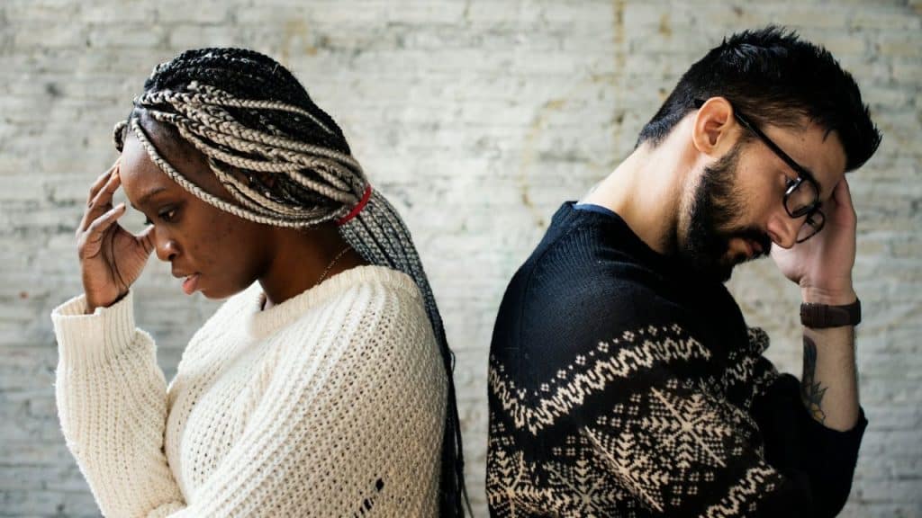 An interracial couple is standing back-to-back, with hands on their heads and looking down, against a white brick wall.
