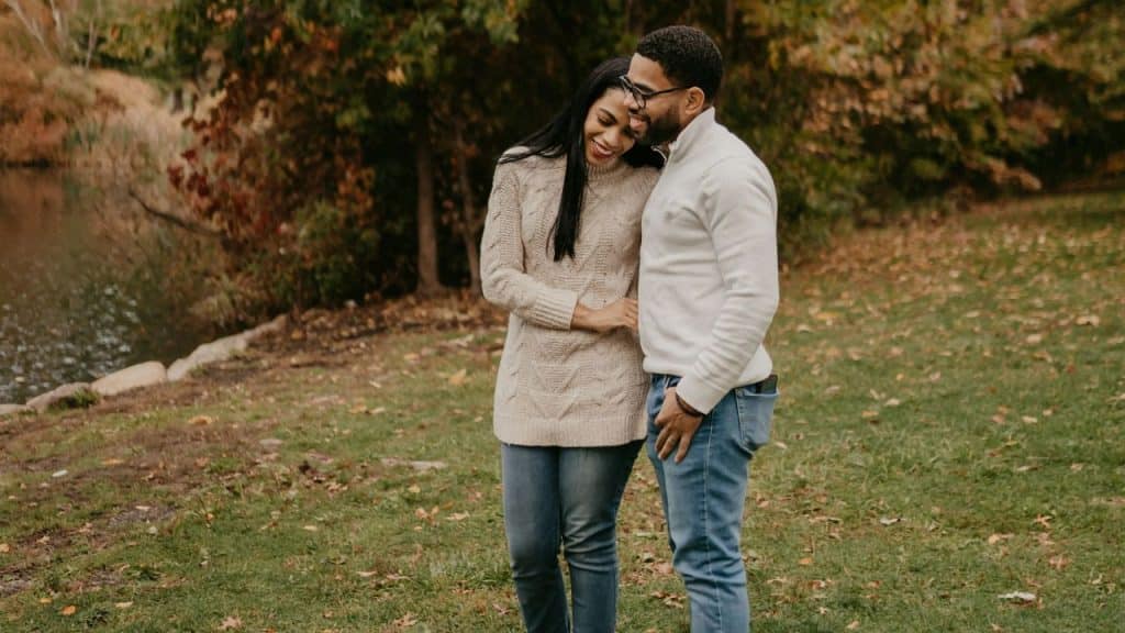 An interracial couple is standing in a park setting near a pond with fall foliage, with the woman leaning her head on the man's shoulder and both smiling.