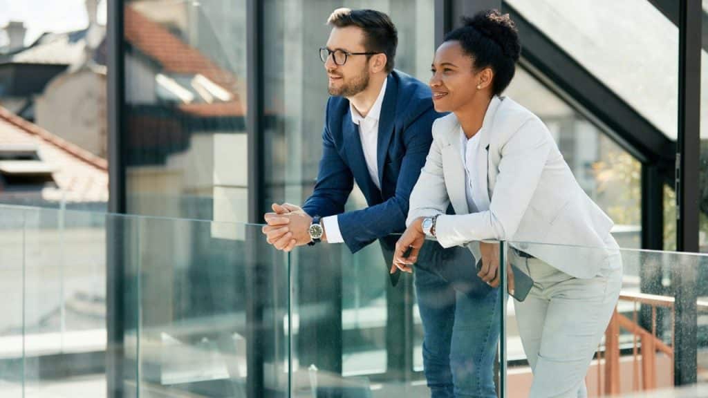 An interracial man and woman in business attire are standing on a balcony, leaning on a glass railing and looking away from the camera.