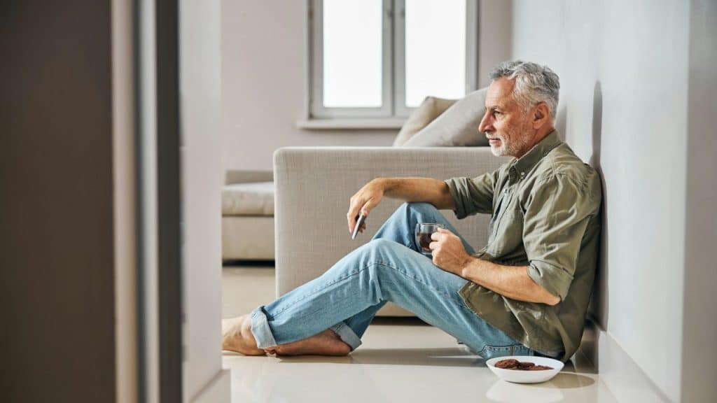 A gray-haired man sits on the floor leaning against a wall, holding a drink and looking away.