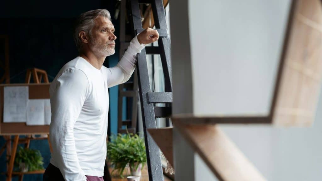 A silver-haired man in a white shirt is standing by a window, looking out thoughtfully.