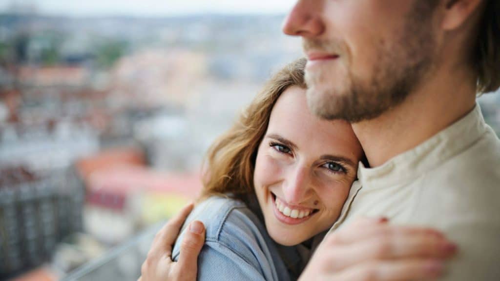A smiling woman is embracing a man on a balcony overlooking a cityscape.
