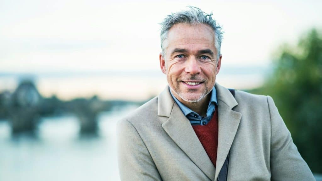 A silver-haired, smiling man in a beige coat stands outdoors with a bridge in the background.