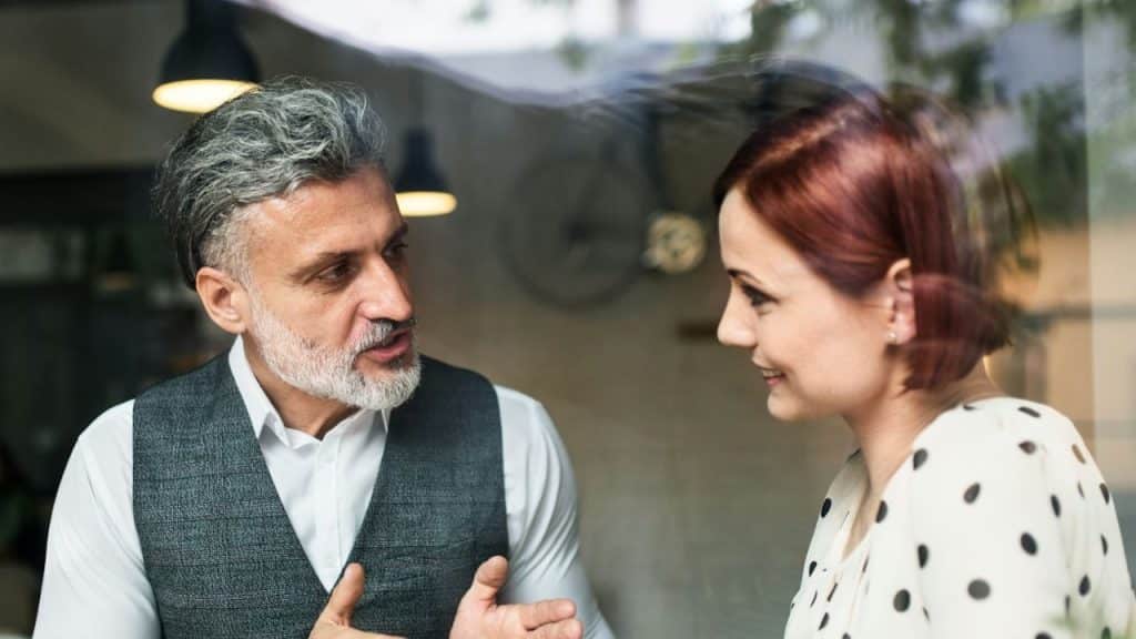 A middle-aged man with a gray beard and vest is talking to a red-haired woman.