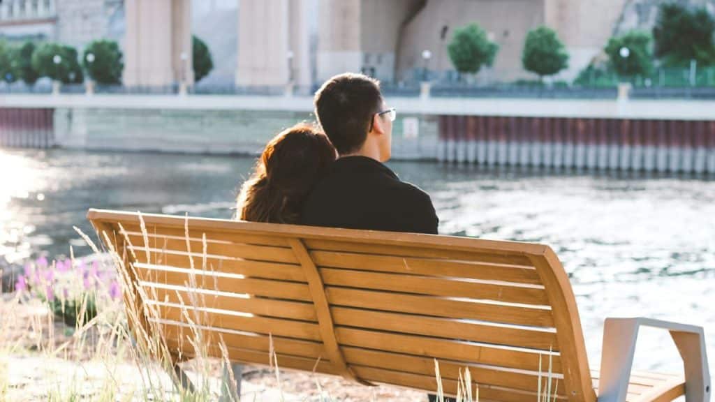 A man and a woman are sitting on a wooden bench by a river looking at the city.