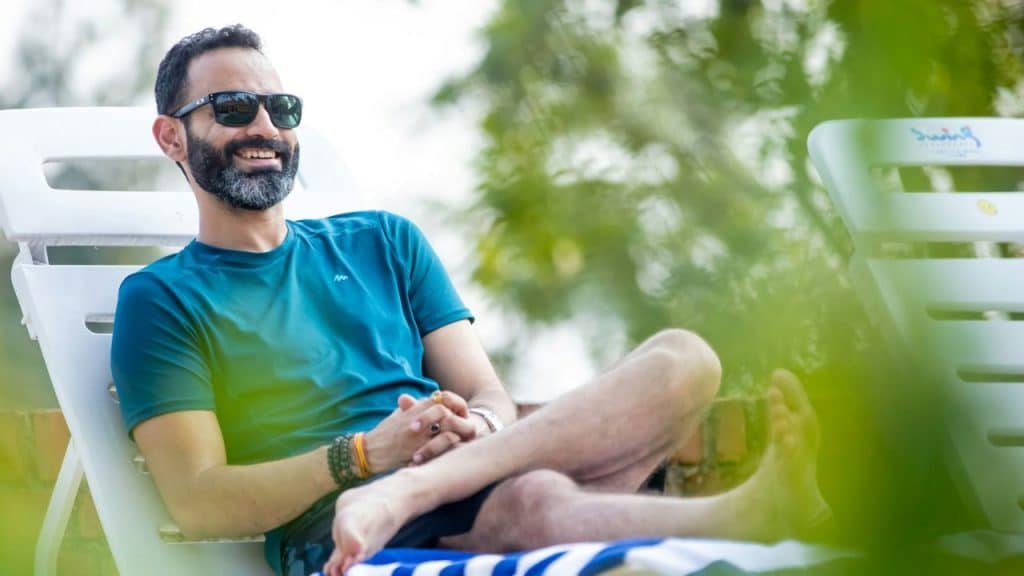 A happy bearded man in a blue shirt and sunglasses is relaxing on a white lounge chair outdoors.